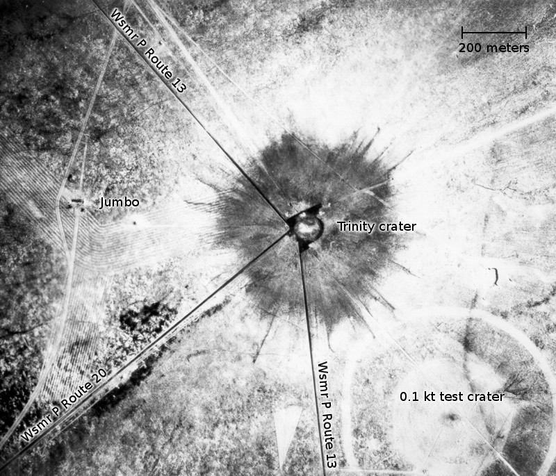 Вид с воздуха на последствия испытания Тринити (атомной бомбы «Штучка») через 28 часов после взрыва. Меньший кратер к юго-востоку образовался от более раннего взрыва 100 тонн тротила 7 мая 1945 года. Слева от кратера виден невредимый контейнер «Джамбо» и его разрушенная башня (вертикальная линия). 18 июля 1945 г.
