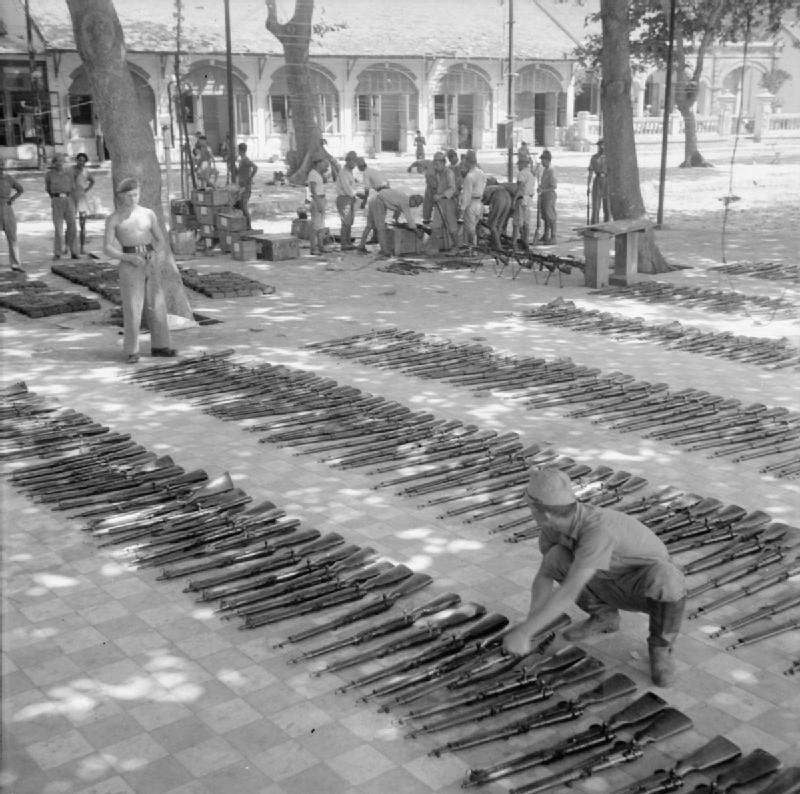 Британские офицеры наблюдают за японскими военнопленными, которым поручено чистить и смазывать тысячи сданного оружия в лагере для военнопленных на мысе Сен-Жак. 1945 г. 
