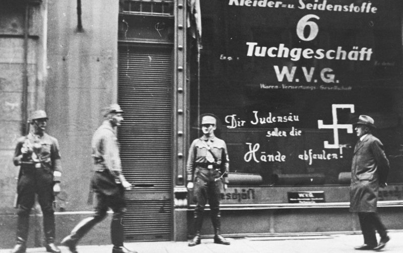 Нацистские штурмовики охраняют принадлежащий евреям магазин в Вене после аншлюса. Надпись на витрине магазина гласит: «Ты, еврейская свинья, да сгниют твои руки!». 1938 г.