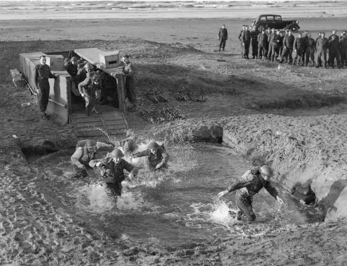 Коммандос имитируют высадку десанта, высаживаясь из макета десантного судна в неглубокую яму, заполненную водой.